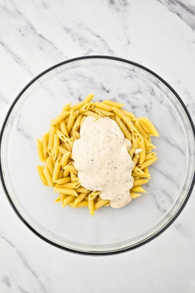 Cooked penne pasta in a glass mixing bowl topped with a generous spoonful of creamy Caesar dressing, showing the second step of preparing a simple cold pasta salad.