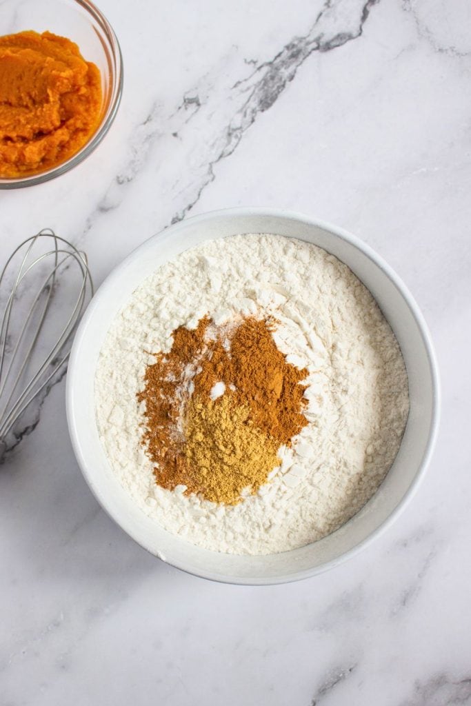 Bowl of flour topped with fall spices including cinnamon, ginger, and baking soda, ready to be mixed. A whisk and a small bowl of pumpkin purée sit nearby on a marble surface, capturing the start of a cozy pumpkin baking recipe.