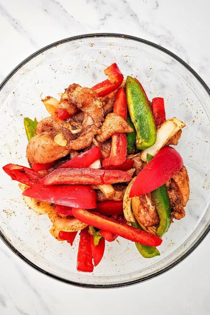 Seasoned chicken strips tossed with sliced red and green bell peppers and onions in a glass mixing bowl.