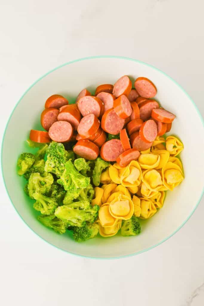 Large mixing bowl filled with sliced smoked sausage, frozen broccoli florets, and cheese tortellini, all prepped for a sheet pan meal. Ingredients are arranged in separate sections, ready to be tossed together for an easy sausage tortellini bake.