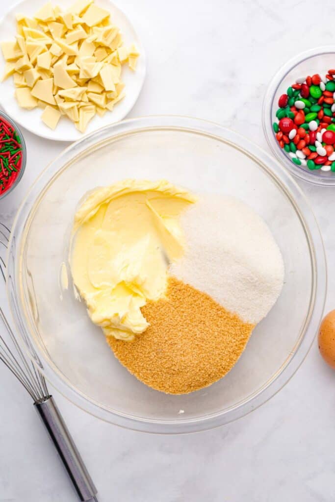 Butter, brown sugar, and white sugar in a large glass mixing bowl, ready to be creamed together, with bowls of chopped white chocolate, Christmas M&Ms, and red and green sprinkles nearby.