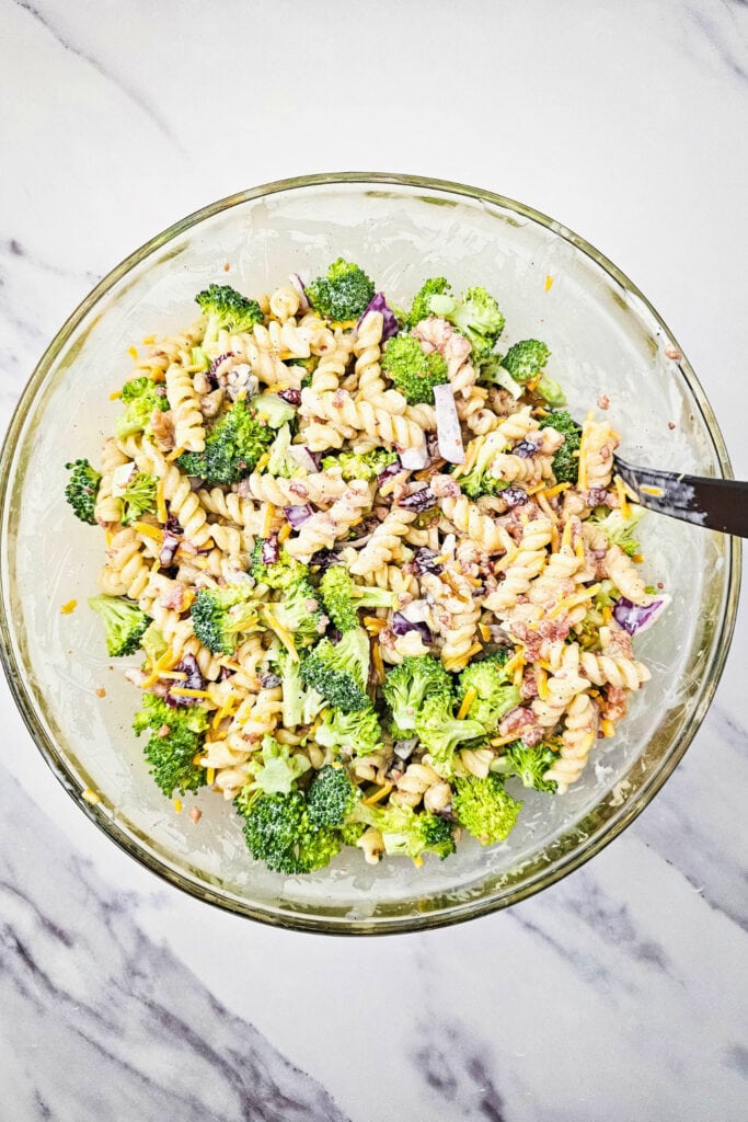 Glass bowl filled with fully mixed broccoli pasta salad, featuring rotini pasta, broccoli florets, red onion, cheddar cheese, dried cranberries, bacon bits, and chopped walnuts coated in a creamy dressing.