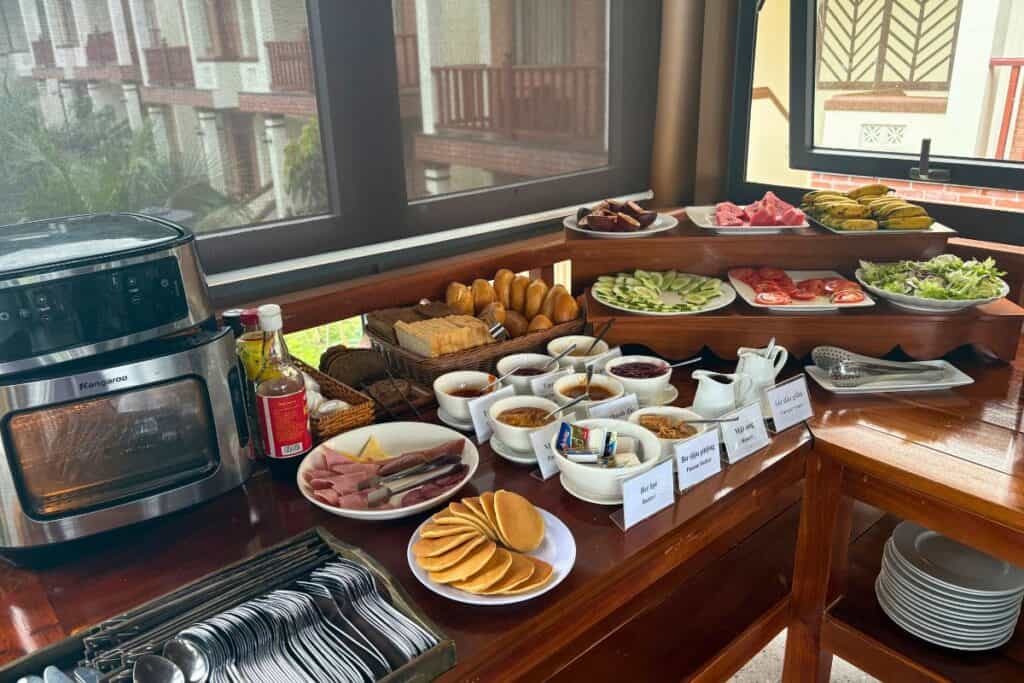 Variety-packed breakfast buffet setup with pancakes, sliced ham, bread, condiments, fresh vegetables, tropical fruits, and sauces arranged neatly on wooden counters. A toaster oven and trays of utensils sit nearby, with natural light coming through screened windows in the background.