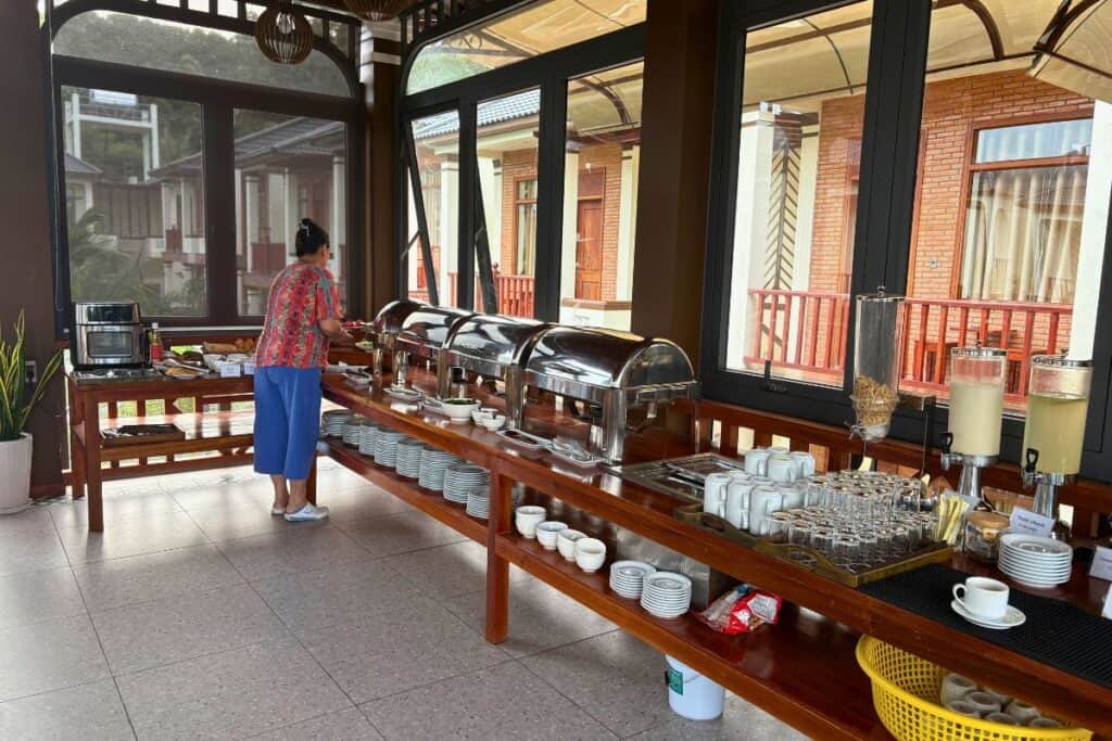 A breakfast buffet setup inside a bright, open dining area with large windows. The buffet includes covered hot dishes, cereal dispensers, juice and milk dispensers, cups, plates, and utensils, while a woman selects food near the counter lined with breads and condiments.