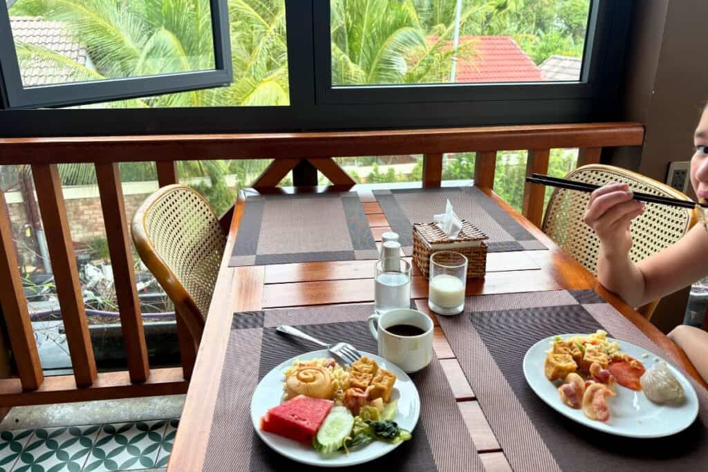 Breakfast at a family resort in Vietnam, featuring plates of tropical fruit, rice, eggs, pastries, and dim sum on a wooden balcony table at Palm Bay Resort in Phu Quoc. A child is seen eating with chopsticks beside a view of lush palm trees through open windows.