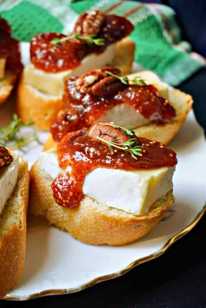Brie and fig jam crostini are arranged on a white serving platter with thick slices of creamy brie resting on toasted baguette rounds. Each crostini is topped with glossy fig jam, pecans, and fresh thyme.