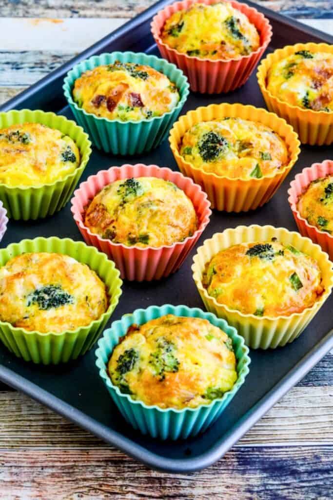 Broccoli bacon cheese breakfast egg muffins baked in colorful silicone cups on a tray with golden tops and visible pieces of broccoli and bacon.