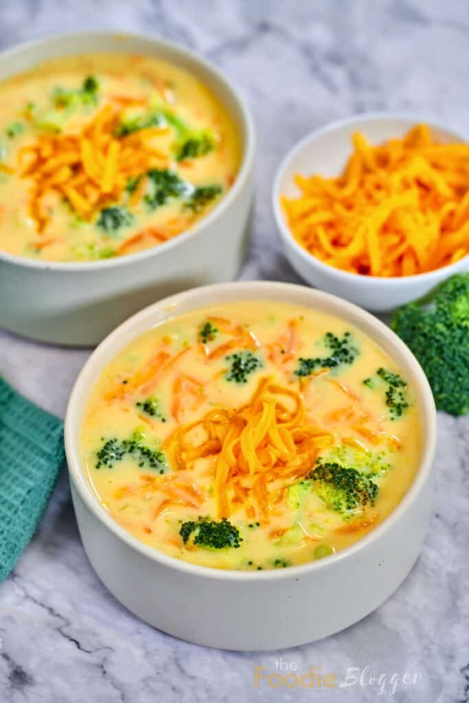 Broccoli cheddar soup with tender broccoli florets and shredded carrots is served in a creamy cheese broth. Shredded cheddar cheese tops the soup with a second bowl in the background.