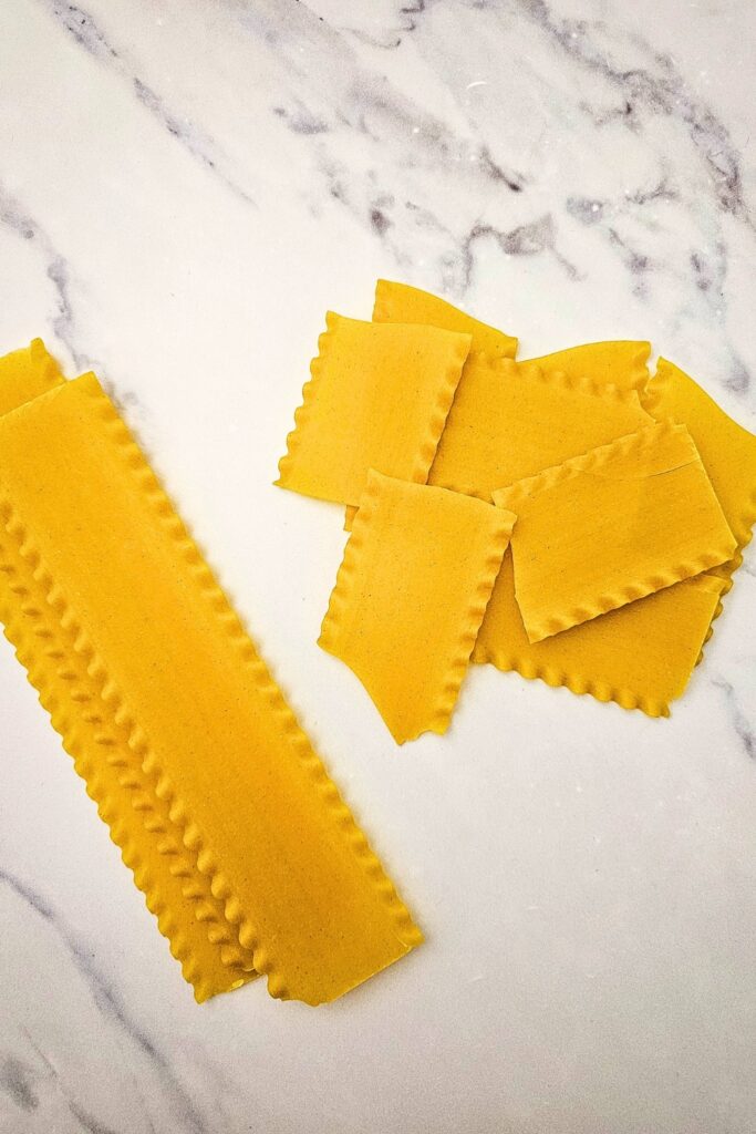 Uncooked lasagna noodles broken into smaller pieces on a marble surface. Breaking the noodles into irregular pieces prepares them for assembling.