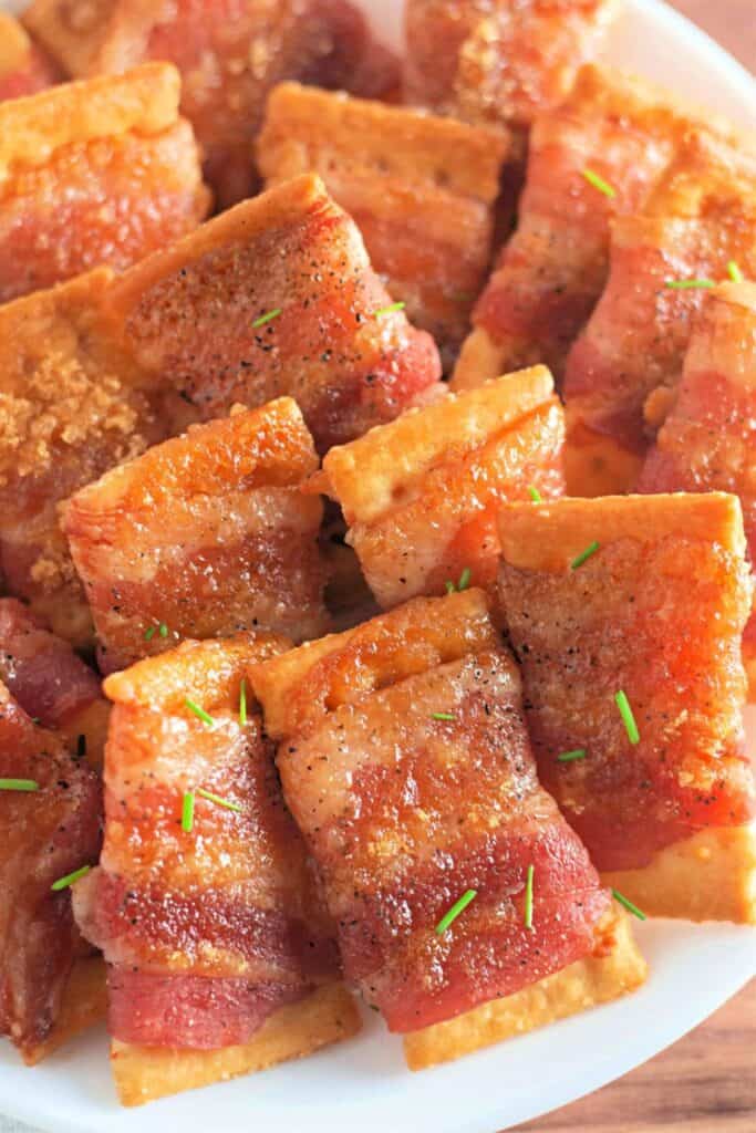 Crispy crackers wrapped in caramelized bacon, sprinkled with brown sugar and black pepper, and garnished with a touch of fresh green herbs, served on a white plate for a savory-sweet snack.