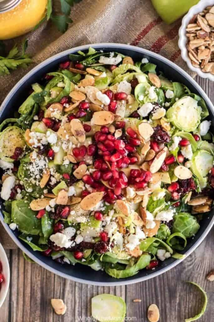 An overhead view of Brussels Sprout Salad with Honey Mustard Vinaigrette made with shaved Brussels sprouts, leafy greens, crisp apple, dried cranberries, crumbled cheese, toasted sliced almonds, and bright red pomegranate seeds.