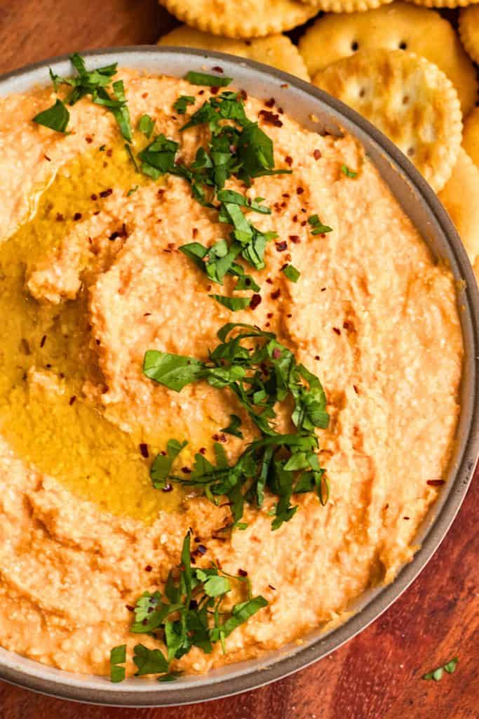 Creamy Buffalo Baked Feta White Bean Dip in a gray bowl, swirled with olive oil and topped with chopped parsley and red pepper flakes. Served alongside round buttery crackers on a wooden surface for a spicy, savory appetizer.