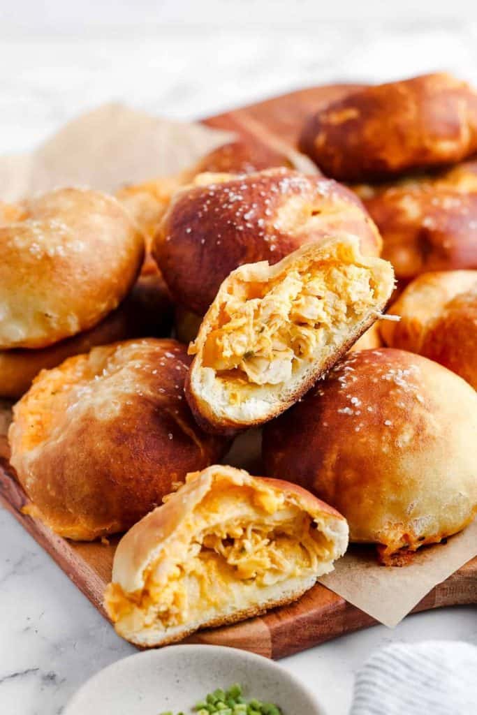 Golden-brown stuffed rolls filled with shredded buffalo chicken and melted cheese, some cut open to reveal the creamy filling. The rolls are sprinkled with coarse salt and arranged on a wooden board lined with parchment paper.