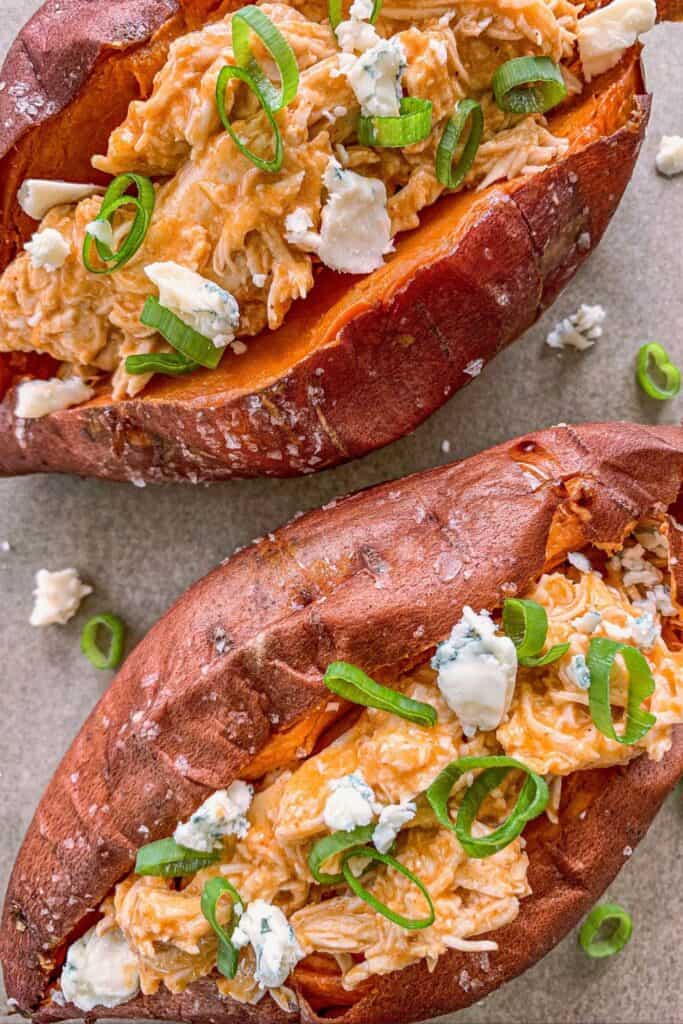 Two baked sweet potatoes topped with shredded buffalo chicken, pieces of blue cheese and green onions.