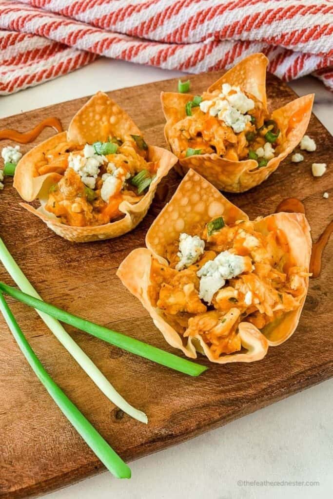 Crispy wonton cups filled with creamy buffalo chicken, crumbled blue cheese, and chopped green onions, served on a wooden board with fresh green onions—perfect finger foods for a football party.