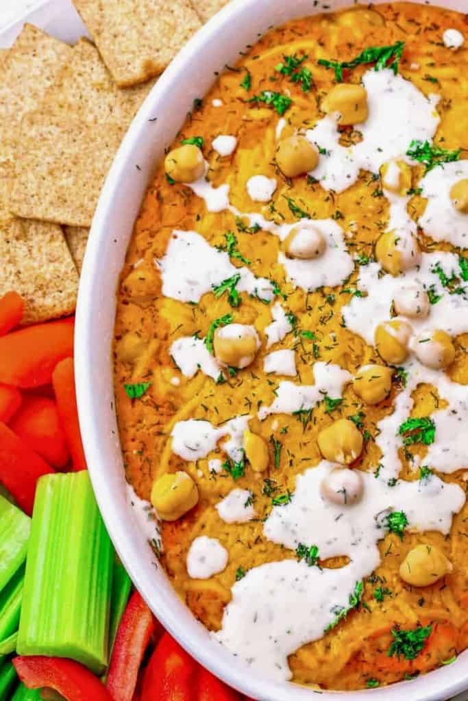 Chilled Buffalo chickpea dip in a white dish, topped with whole chickpeas, a drizzle of creamy ranch dressing, and a sprinkle of fresh herbs. A cold game day dip surrounded by whole grain crackers, celery sticks, and sliced red bell peppers.