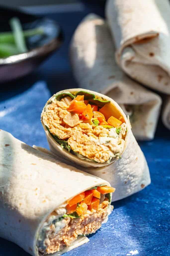 Buffalo chickpea wraps sliced in half to reveal a creamy buffalo chickpea filling with chopped carrots and leafy greens inside a soft tortilla. The wraps rest on a blue surface with extra rolled tortillas in the background.