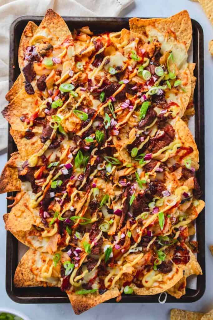 A sheet pan filled with bulgogi and kimchi nachos, layered with tortilla chips, melted cheese, marinated Korean beef, sautéed kimchi, and drizzled with spicy mayo and barbecue sauce, topped with chopped red onions and sliced green onions for a bold, savory twist on classic nachos.