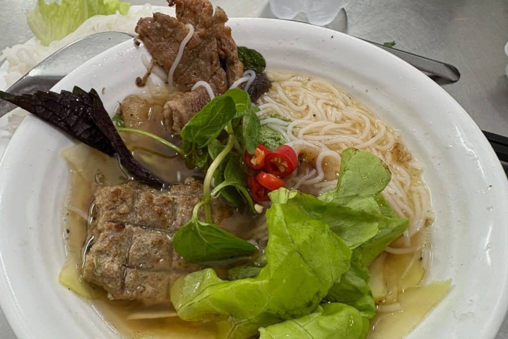 A bowl of bún chả features grilled pork, minced pork patties, vermicelli noodles, fresh herbs, lettuce, and sliced red chili in a light, flavorful broth. The dish is served with a spoon and chopsticks on the side, showcasing the vibrant and balanced ingredients typical of this Vietnamese specialty.