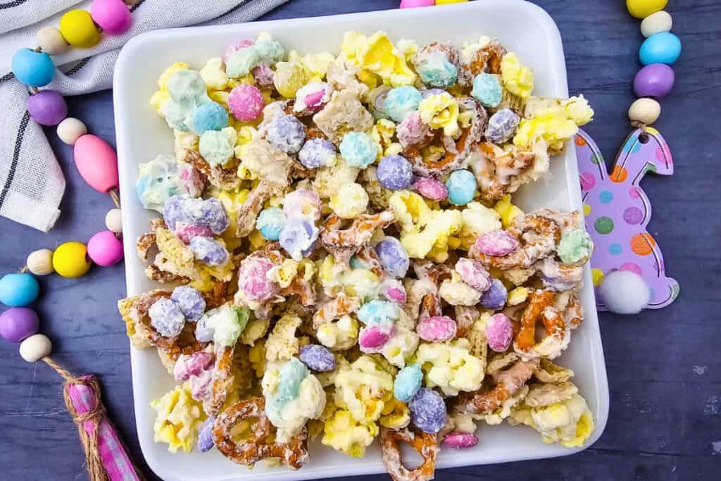 Colorful Easter snack mix with popcorn, pretzels, and pastel candy pieces coated in white chocolate, served in a white bowl surrounded by bright spring decorations.