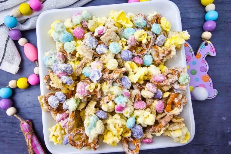 Colorful Easter snack mix with popcorn, pretzels, and pastel candy pieces coated in white chocolate, served in a white bowl surrounded by bright spring decorations.