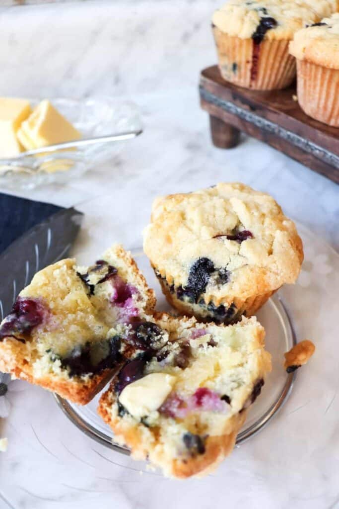 Buttermilk blueberry muffins with a golden crumb topping and juicy blueberries baked inside shown split open with butter melting into the soft interior. One whole muffin sits beside the halves while more muffins rest in the background.