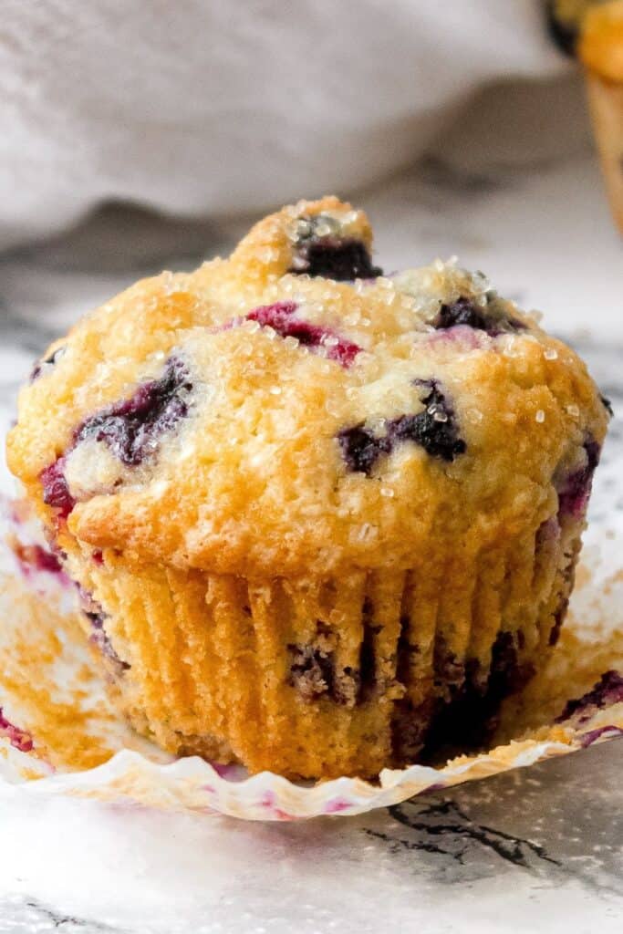 Close up of a buttermilk blueberry muffin with a golden brown domed top sprinkled with coarse sugar crystals. Juicy blueberries burst through the tender crumb and the paper liner is peeled back to show the moist texture.