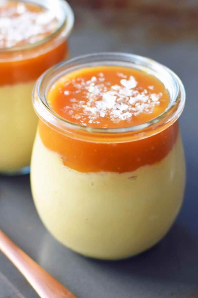 Butterscotch Budino with Salted Caramel served in small glass jars with creamy golden custard beneath a thick layer of caramel sauce. Flaky sea salt is sprinkled over the caramel for a sweet and salty finish.