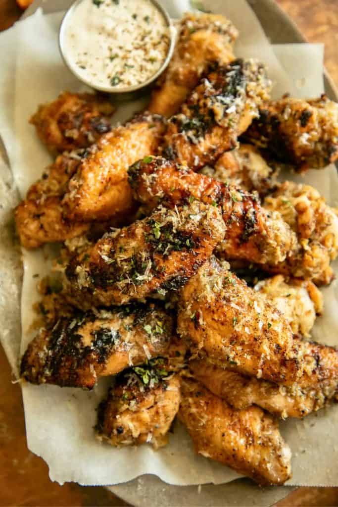 Pile of grilled chicken wings seasoned with herbs and grated parmesan, served on parchment paper with a small metal cup of creamy dipping sauce. The wings have a crispy, golden-brown exterior with visible char marks and flecks of seasoning.