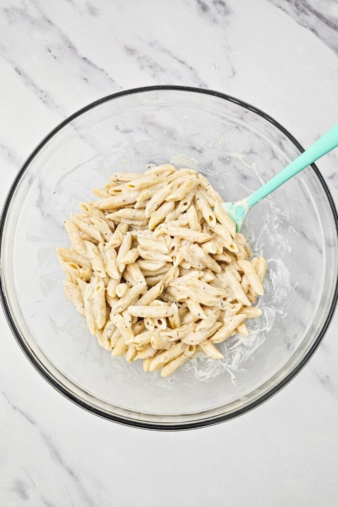 Cooked penne pasta coated in creamy Caesar dressing being mixed in a glass bowl with a spoon, showing the step of tossing pasta for a cold penne pasta dish.