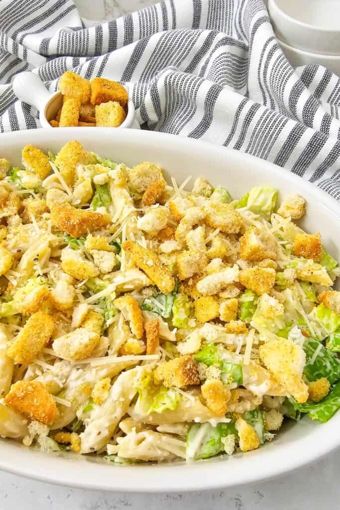 Caesar salad with pasta served in a white dish, featuring penne coated in creamy dressing, crisp romaine lettuce, shaved Parmesan, and crunchy croutons, styled as a hearty Caesar salad with pasta ready to serve.