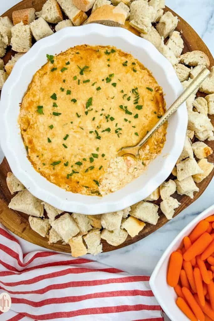 Golden-baked Cajun Crab Party Dip in a white dish, topped with fresh parsley, surrounded by cubed bread and baby carrots for dipping.