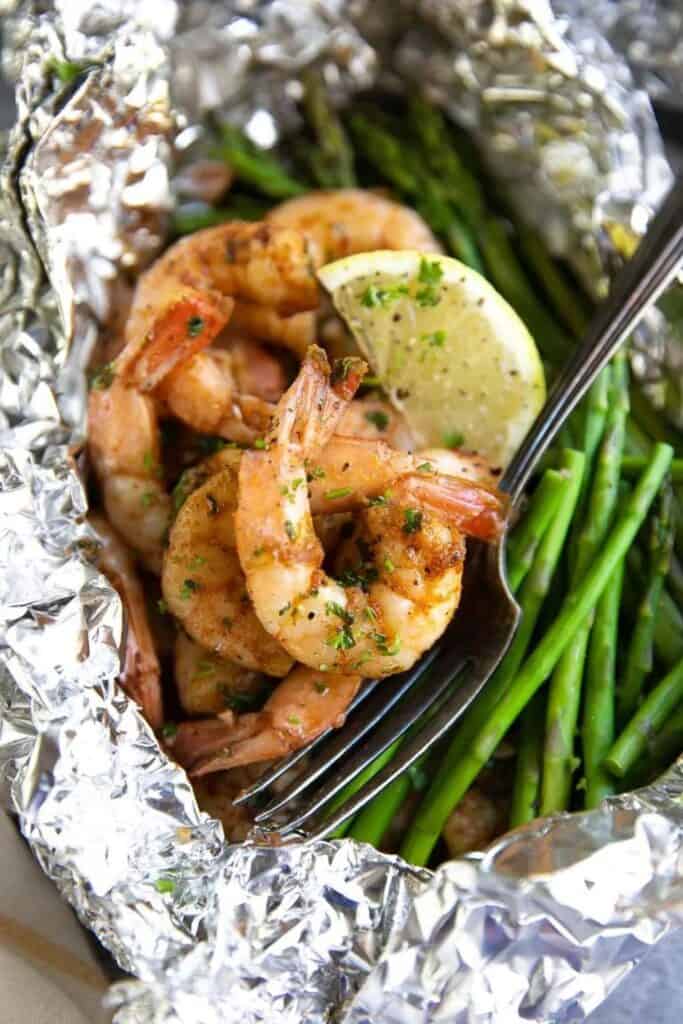 Overhead close up view of an open foil packet with cajun shrimp and asparagus topped with lemon and a fork. 
