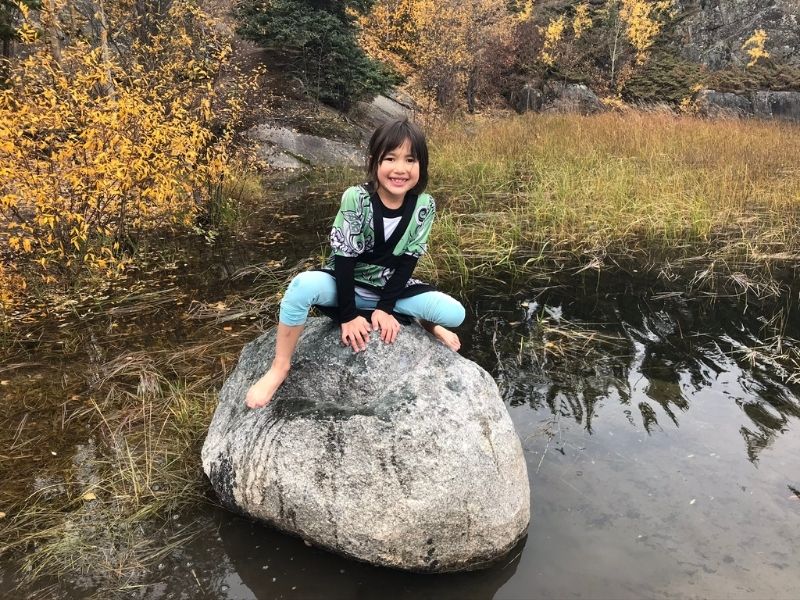 Picture of a young girl sitting barefoot on a rock surrounded by shallow water. Bushes in the background.