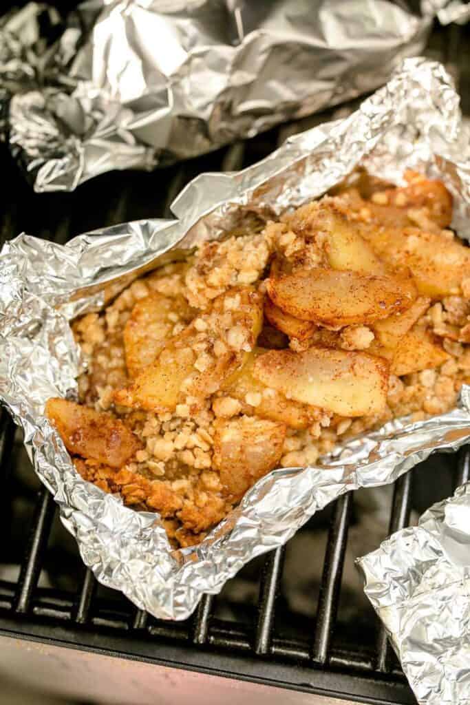 Foil packet filled with cinnamon-sugar apples and crumb topping cooking on a grill grate over a campfire. The tender apple slices are coated in a buttery oat crumble, creating a rustic outdoor apple crisp dessert.