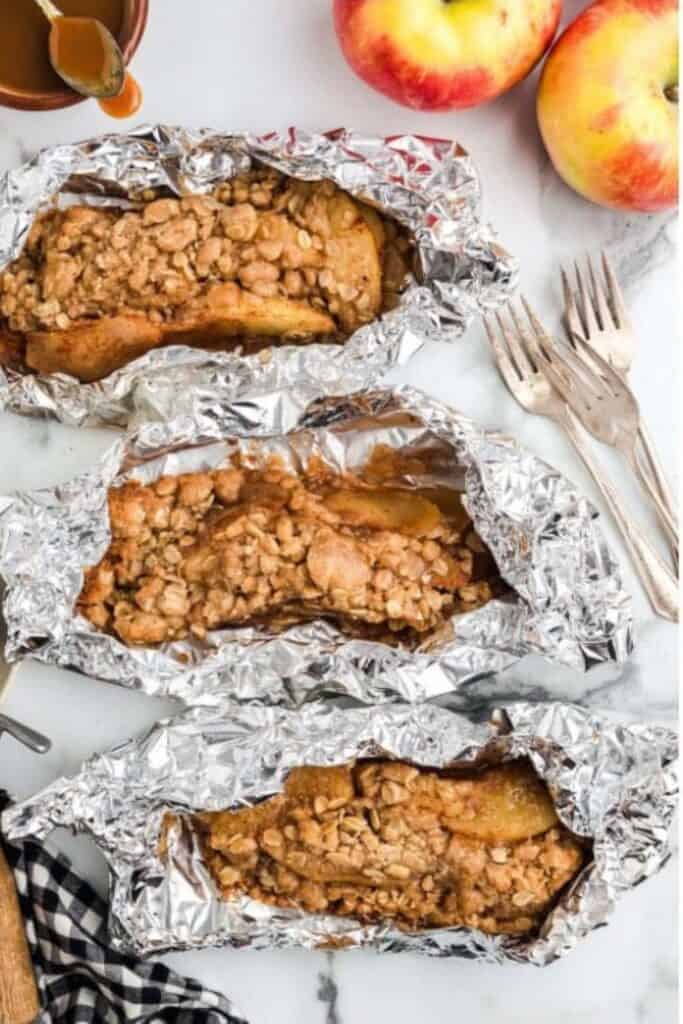 Three foil packets of apple crisp with forks and red apples on a table top.