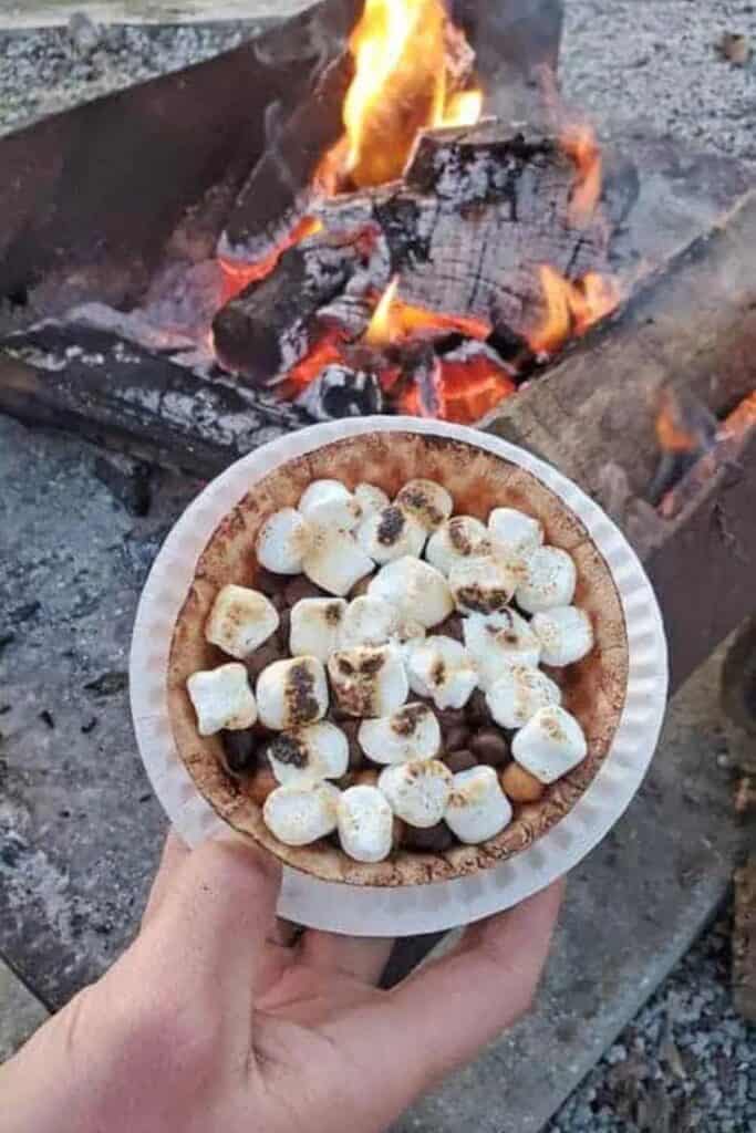 Hand holding a small round tinfoil pan filled with marshmallows, chocolate chips and banana slices in a waffle bowl with a campfire in the background. 
