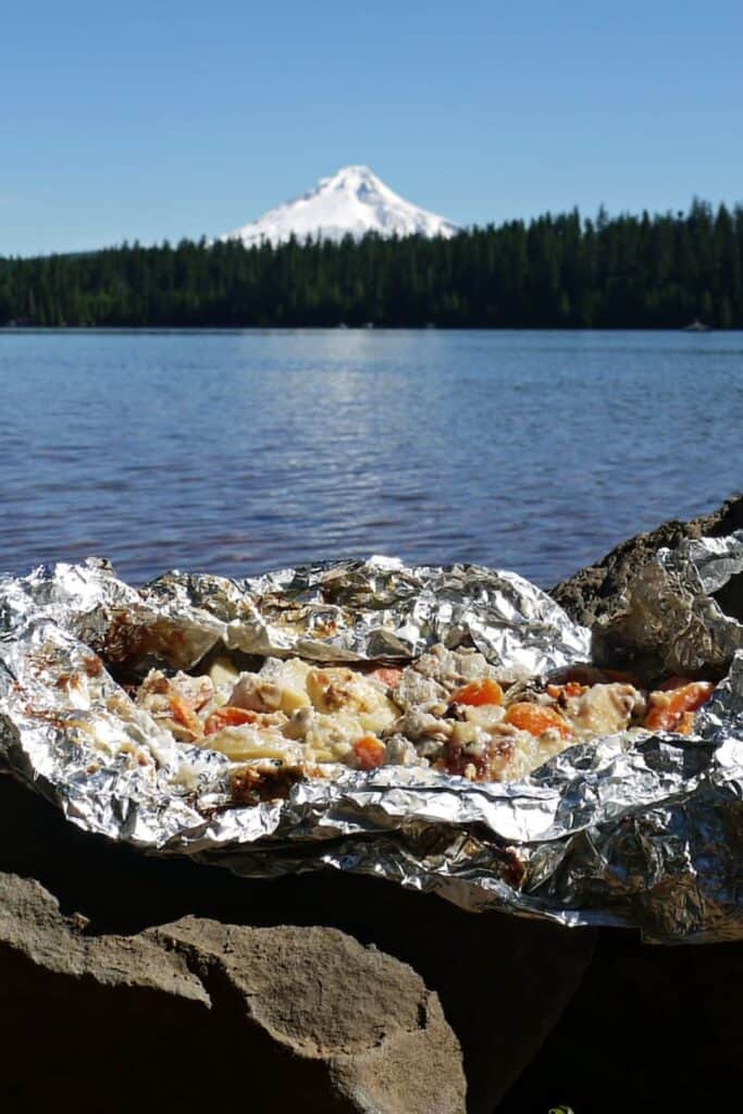 Campfire foil pack stew cooked in crinkled aluminum foil and set on rocks beside a calm lake with a snow capped mountain in the background. The foil packet is opened to reveal tender chunks of meat, potatoes, and carrots coated in melted cheese and savory juices.