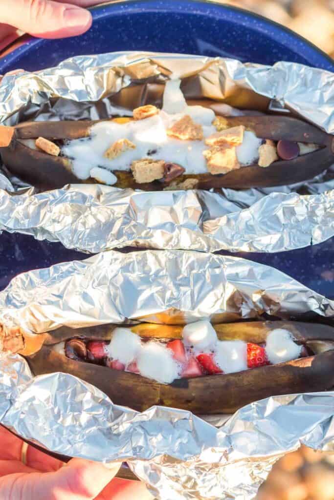 Camping plate with two foil wrapped banana boats topped with marshmallows, strawberries and melted chocolate.