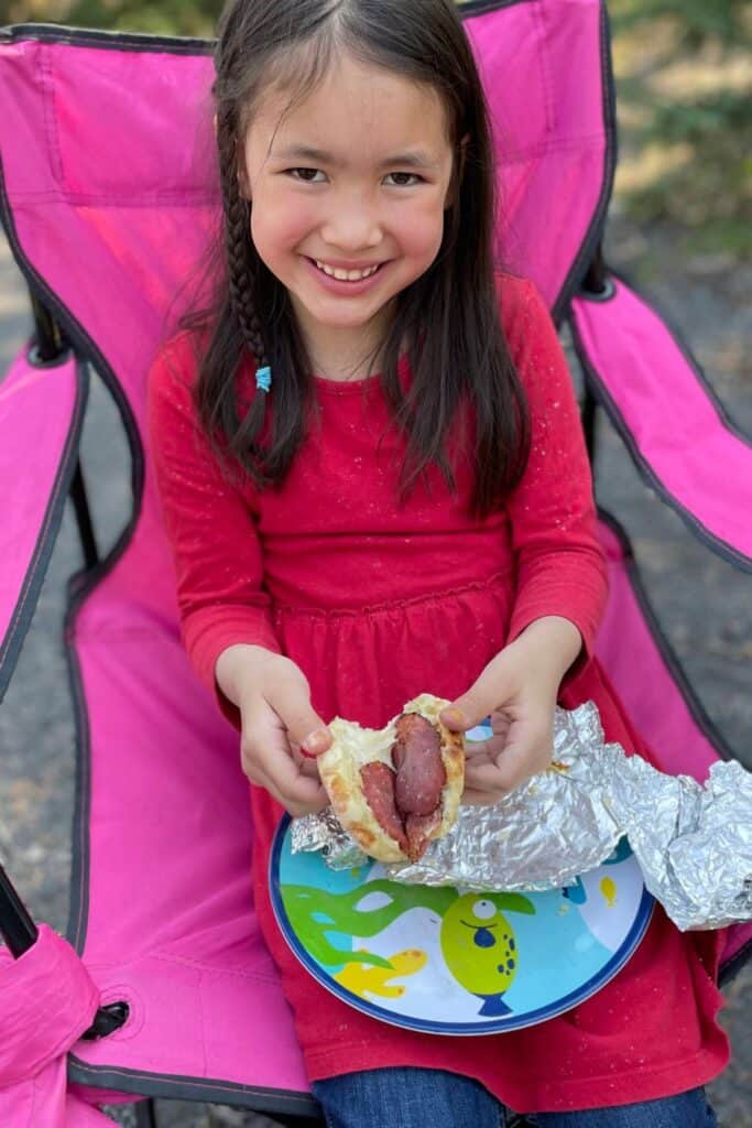 Tween girl sitting in a camp chair, smiling holding a english muffin breakfast sandwich.