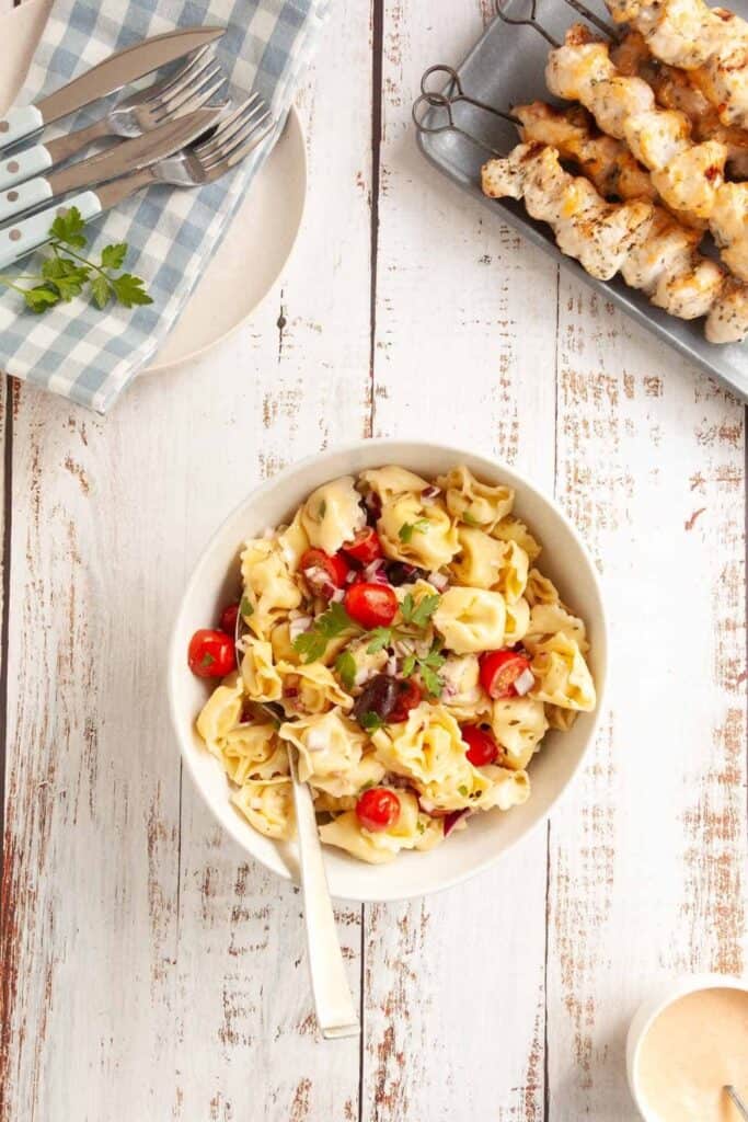Bowl of tortellini camping pasta salad with cherry tomatoes red onion olives and parsley in a creamy dressing on a rustic wooden table. Grilled chicken skewers sit nearby with picnic plates and utensils on a blue gingham cloth showing an easy outdoor campsite meal.