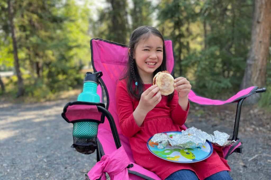 Young girl sitting in a camp chair smiling with a breakfast sandwich in her hands.