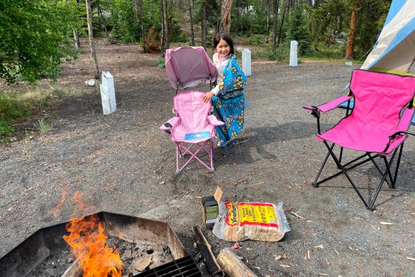 Young girl smiling beside two camp chairs and a campfire.