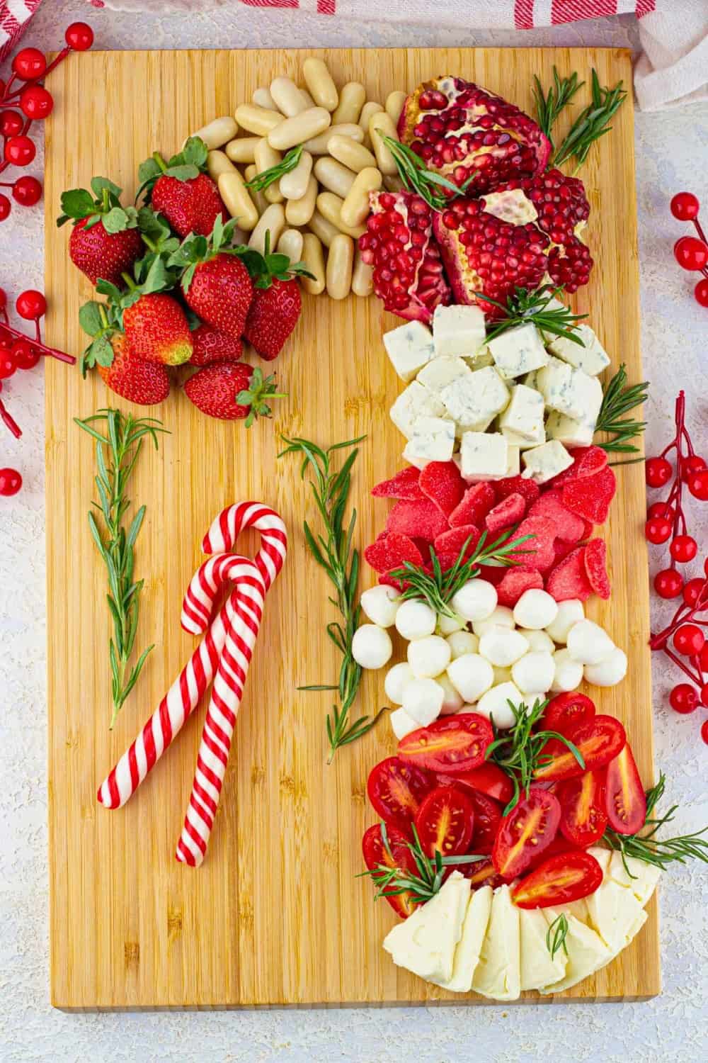 A festive candy cane charcuterie board arranged with strawberries, pomegranate, cheeses, tomatoes, red candy, white candy, and fresh rosemary. Two red and white striped candy canes rest beside the food on the wooden serving board.