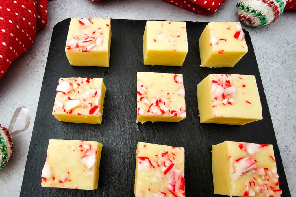Nine square pieces of creamy white fudge topped with crushed peppermint candy arranged neatly on a black slate board. This no-bake candy cane fudge recipe is styled festively with red and white holiday decorations in the background.