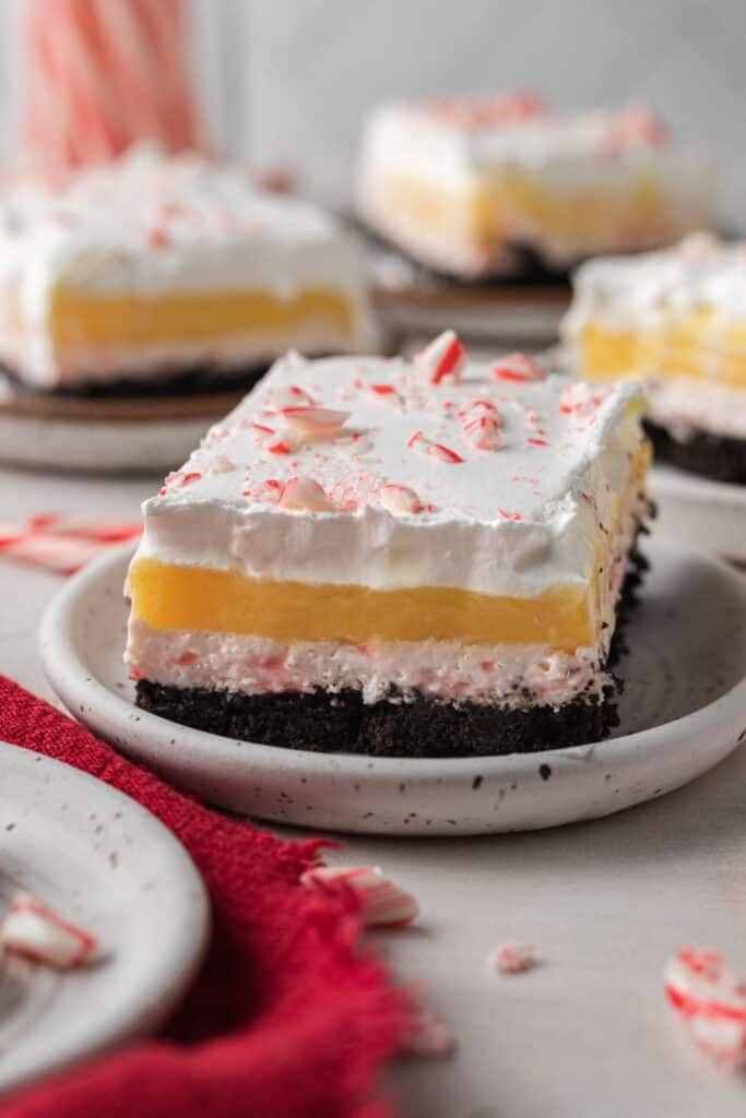 Layered candy cane dessert bar with a chocolate cookie crust, creamy peppermint layer, bright yellow pudding center, and whipped topping sprinkled with crushed candy canes. Served on a ceramic plate with festive red accents and broken peppermint pieces nearby.