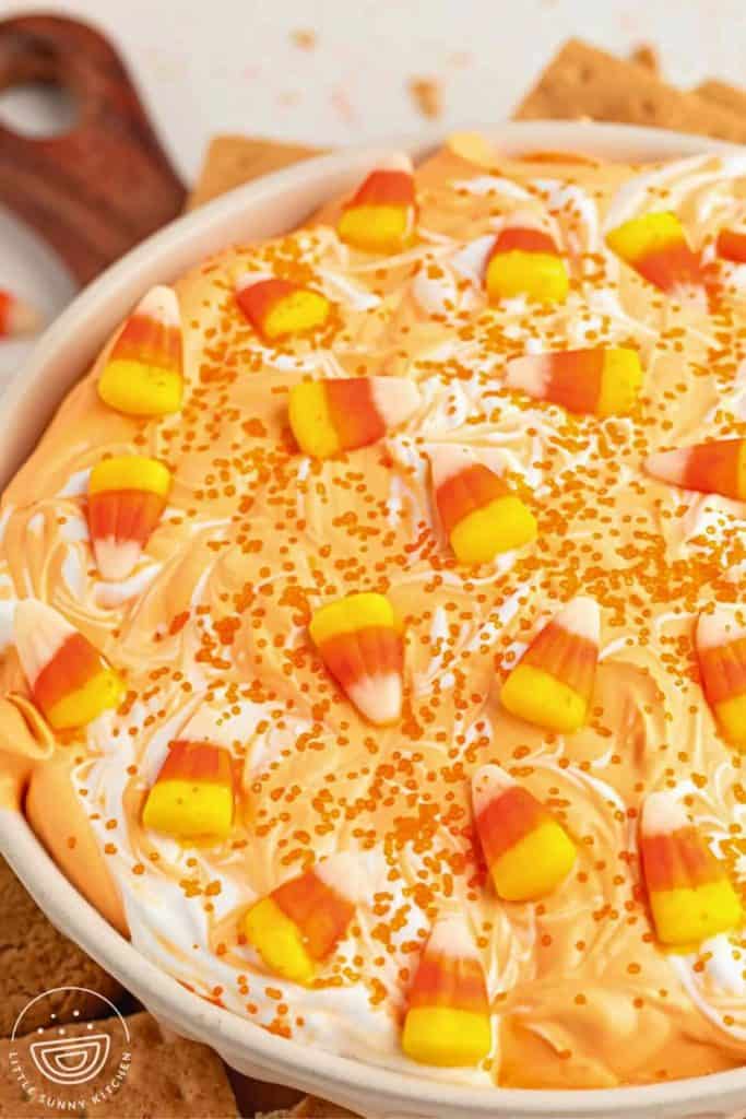 Close-up of a creamy Halloween dessert dip swirled with orange and white colors, topped with candy corn and orange sprinkles. Served in a round dish with graham crackers nearby for dipping.