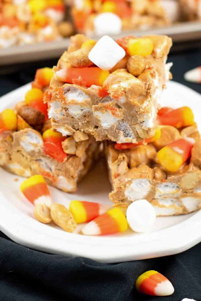 Close-up of Halloween candy bars made with layers of candy corn, mini marshmallows, peanuts, and a creamy peanut butter mixture, cut into squares and stacked on a white plate. A festive Halloween treat with a chewy, crunchy texture and colorful seasonal candy.