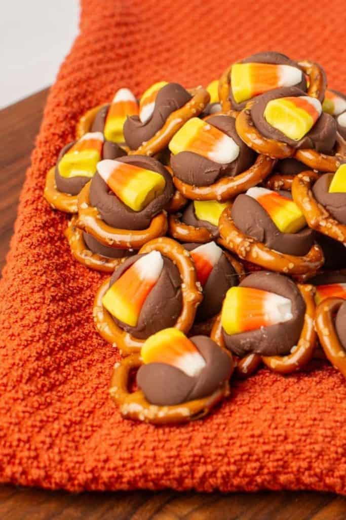 Pile of Candy Corn Pretzel Hugs resting on a textured orange towel, made with mini pretzels topped with melted chocolate hugs and candy corn. The sweet and salty Halloween treat pops with festive fall colors.