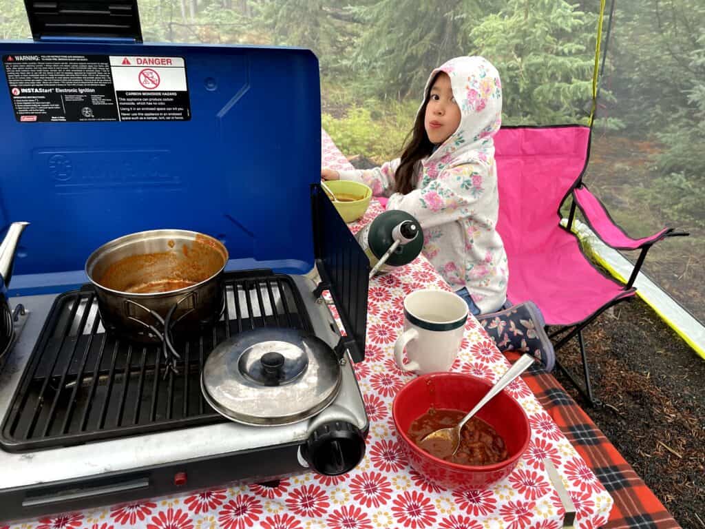 Best canned foods for camping, chili for breakfast. Camp stove and young girl with bowls of chili. 